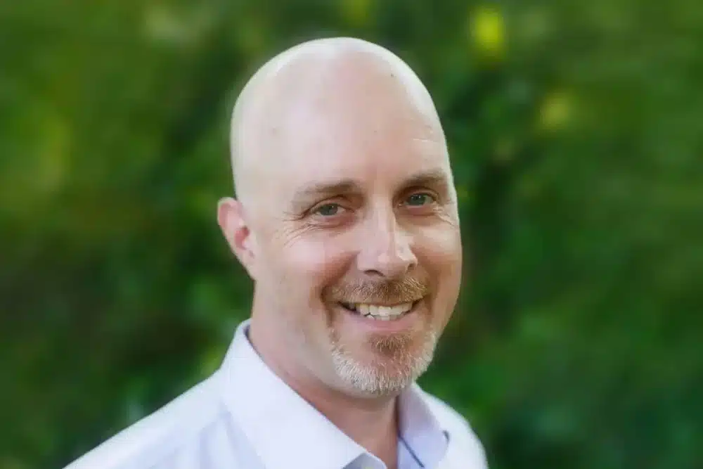 damian-bushnell A man with a bald head and a light beard is smiling at the camera. He is wearing a light-colored shirt and stands against a blurred, green outdoor background.