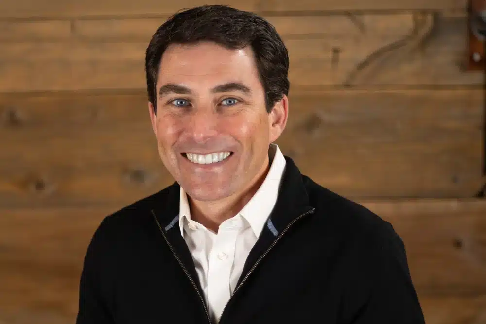 bryan-sullivan A smiling man with short dark hair is wearing a white shirt under a black jacket. He is standing in front of a wooden background.