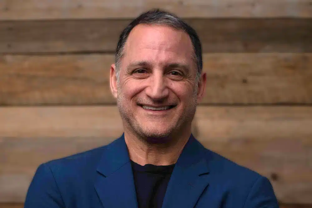 jeff-mandel A man with graying hair and stubble smiles warmly at the camera. He is wearing a dark blue blazer over a black shirt and stands in front of a wooden plank background.